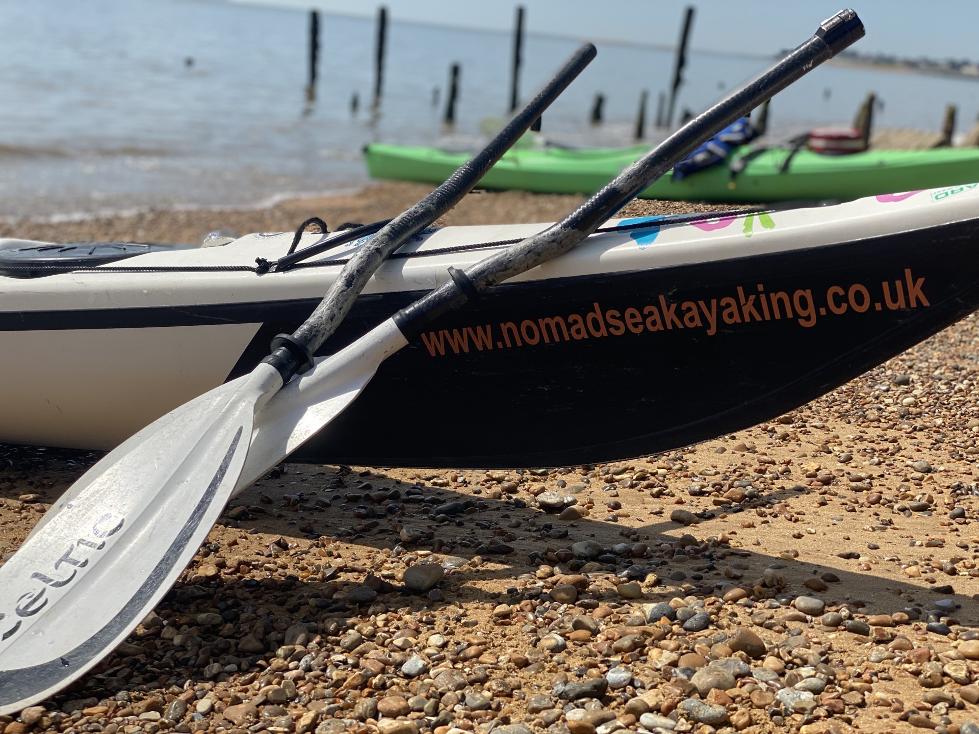 A white Celtic paddle split in two & resting on a sea kayak with another sea kayak in the background
