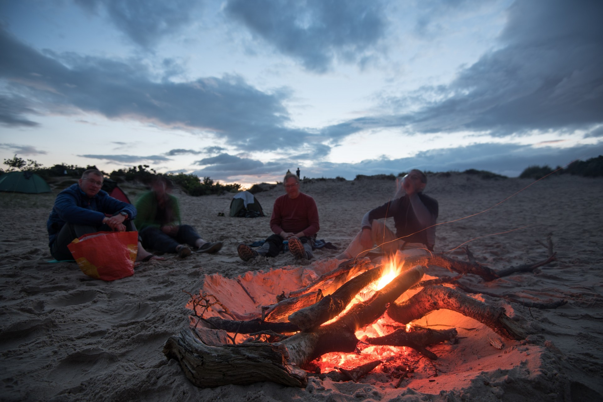 Four people sitting around campfire on beach at dusk, wild camp and kayaking