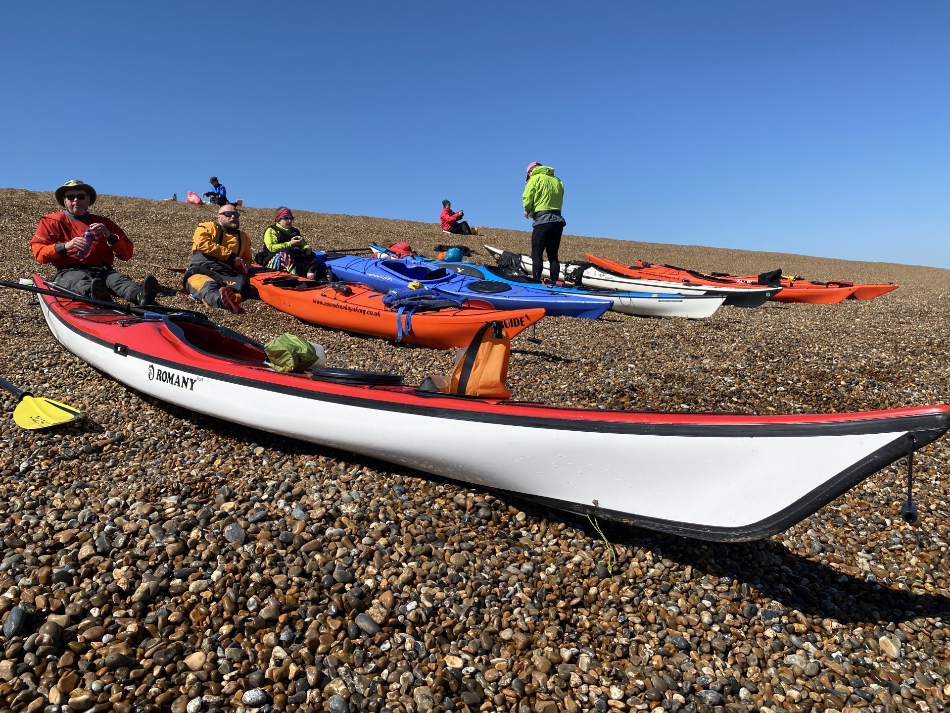 Kayakers taking a break on a shingle beach with bright blue skies.