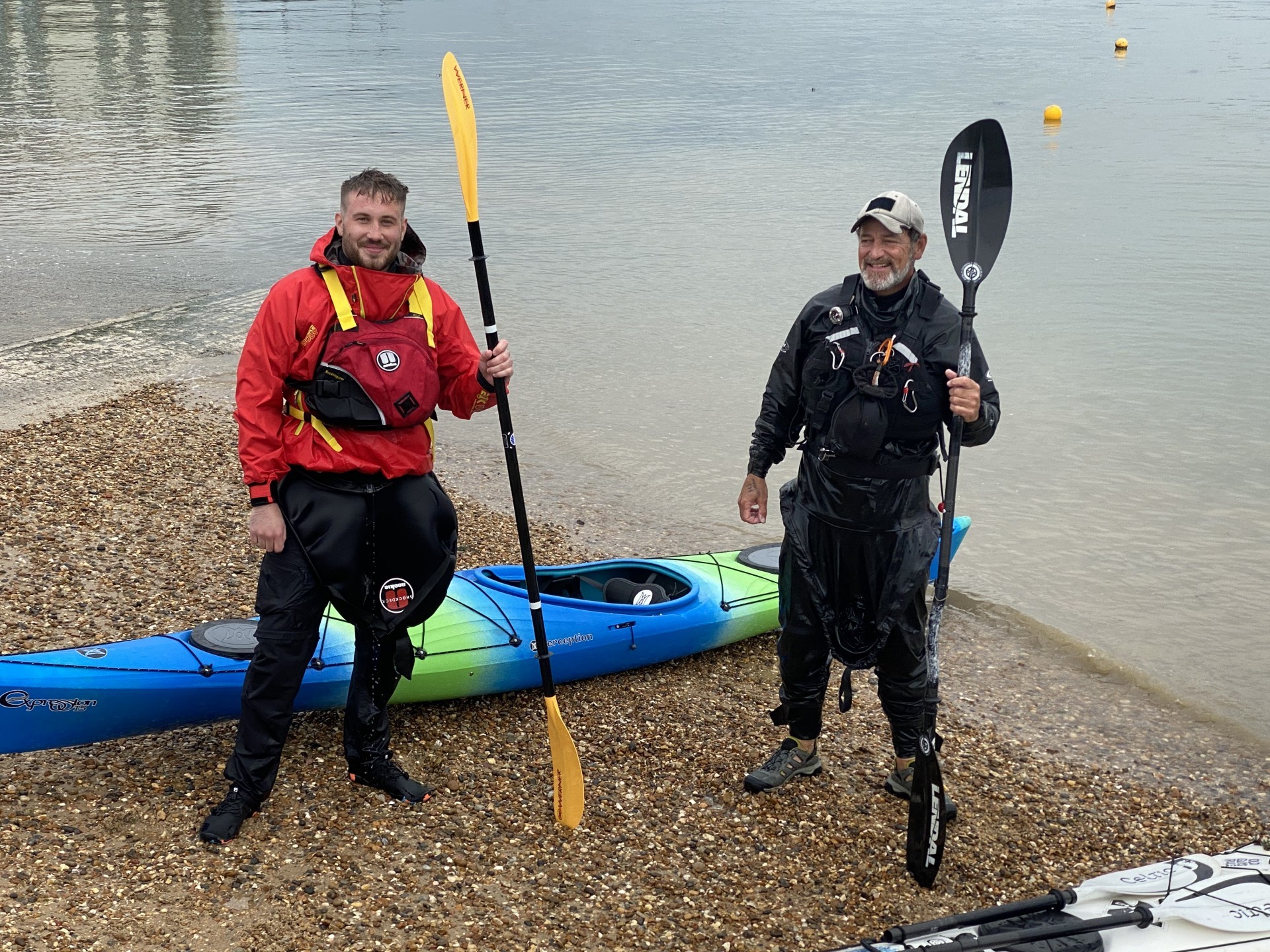 Two smiling kayakers on a beach with their paddles & kayaks