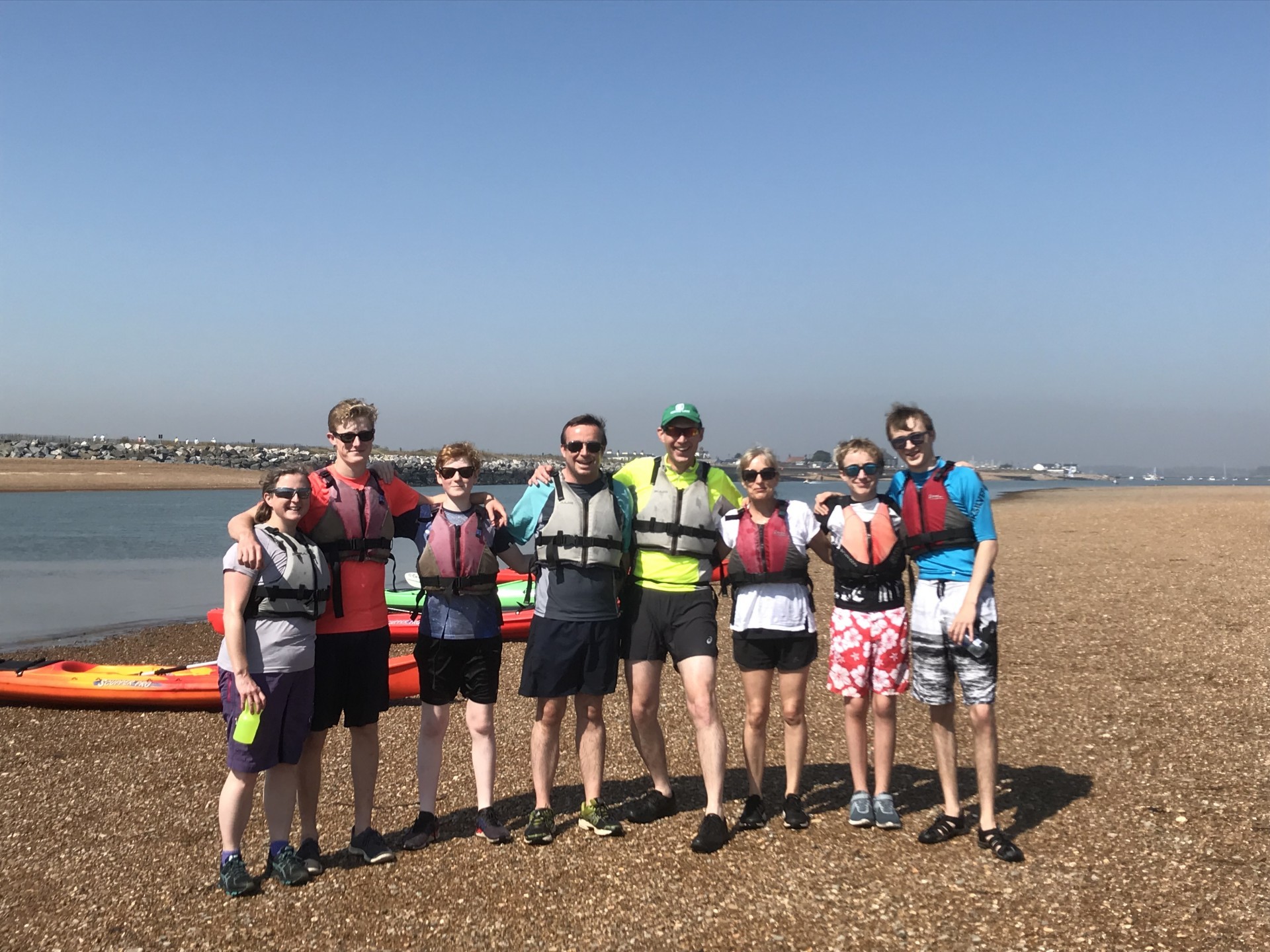 A large family of adults, teenagers and youngsters posing together on a shingle shoal with kayaks in the background