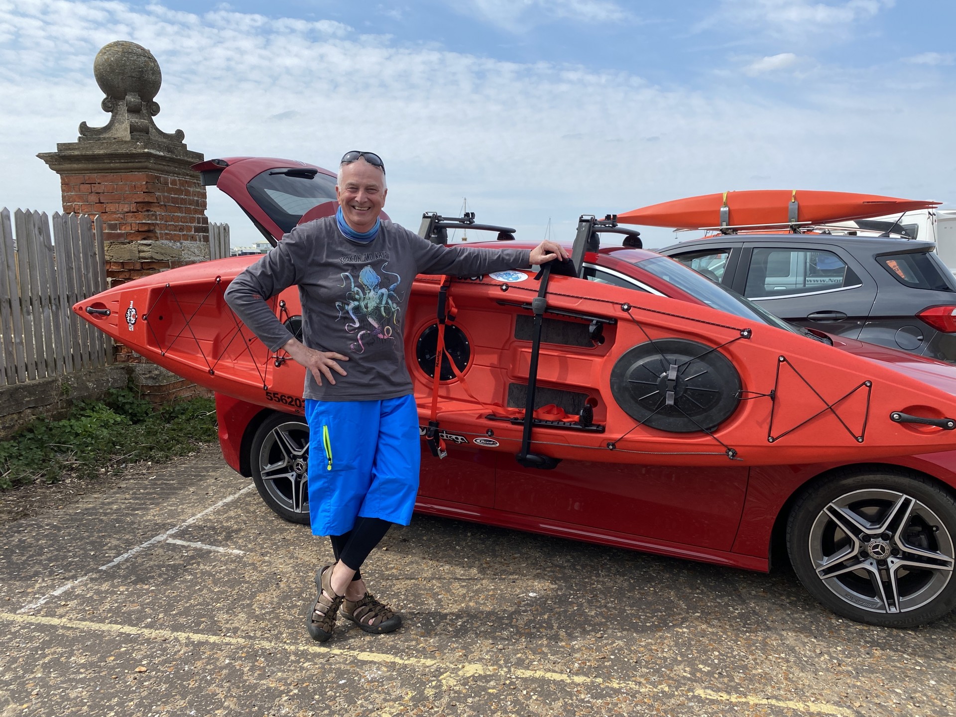 A kayaker standing in front of his kayak wearing a long sleeve 'Krak'on and Kayak' NSK shirt.