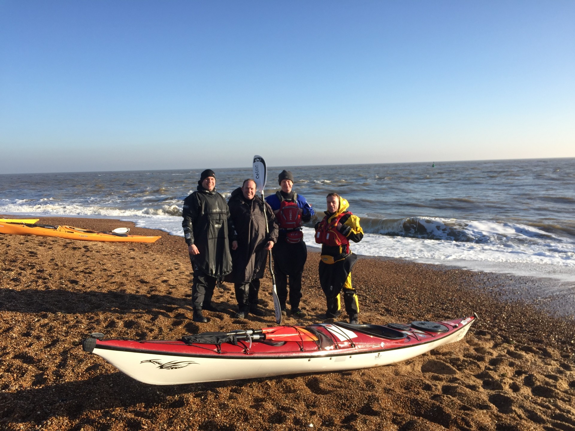 Group os sea kayakers posing together on a shingle beach with clear blue skies