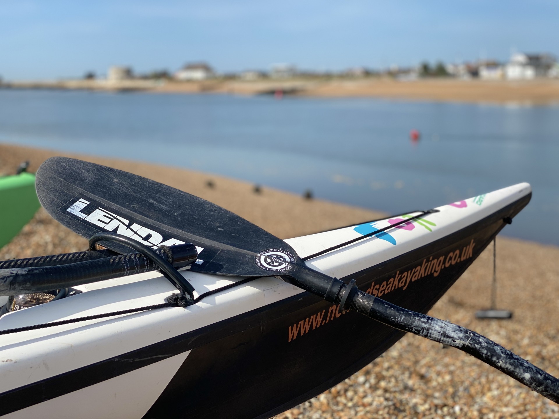 A black carbon fibre Lendal paddle resting against a sea kayak on a beautiful shingle beach