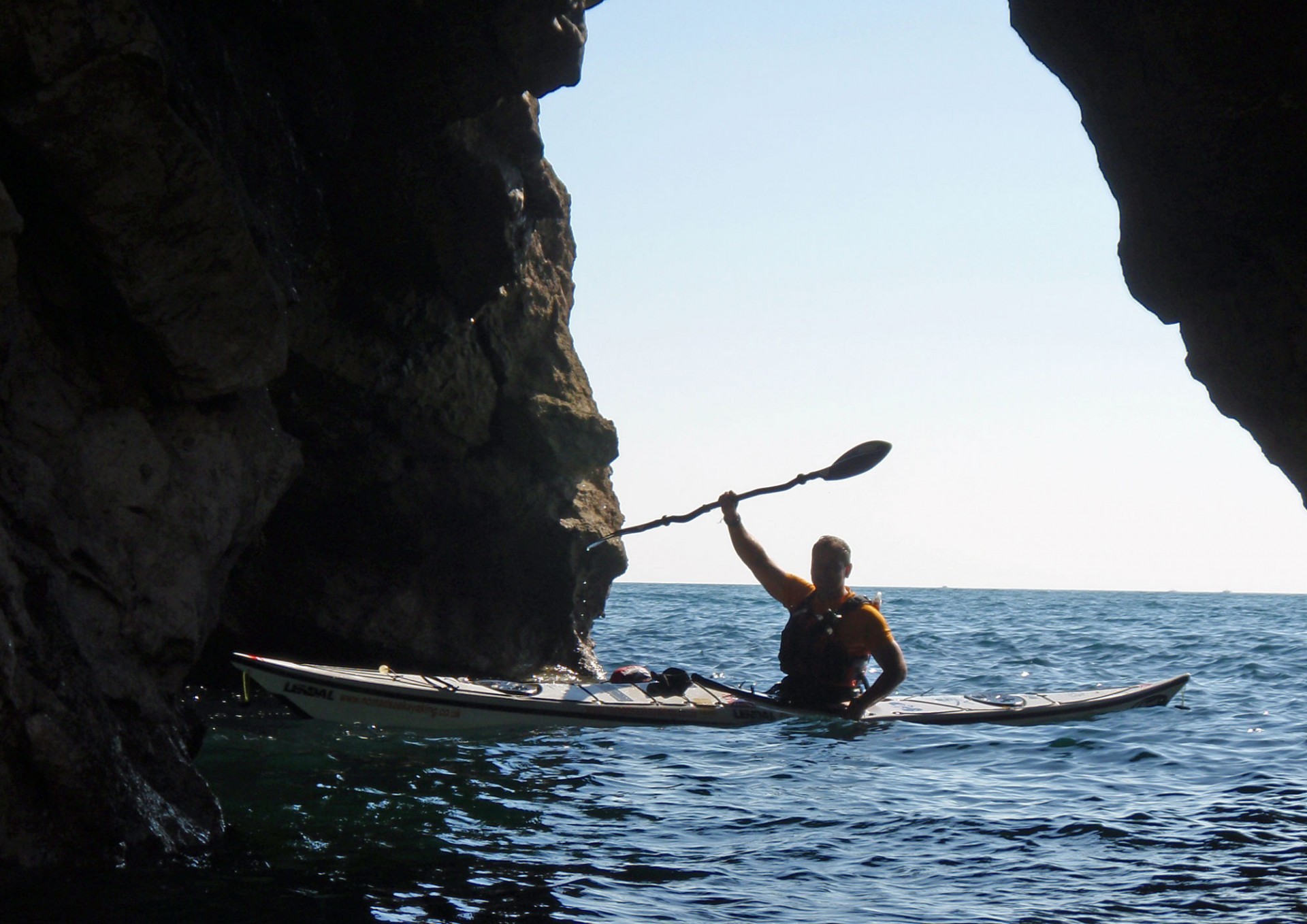 Sea kayaker in a sea cave triumphantly holding a paddle aloft